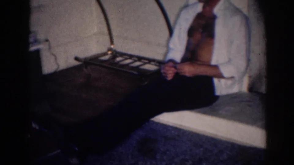 1967 Los Angeles 8mm frame of an adult man in an open white shirt seated beside a metal luggage rack in a private interior.