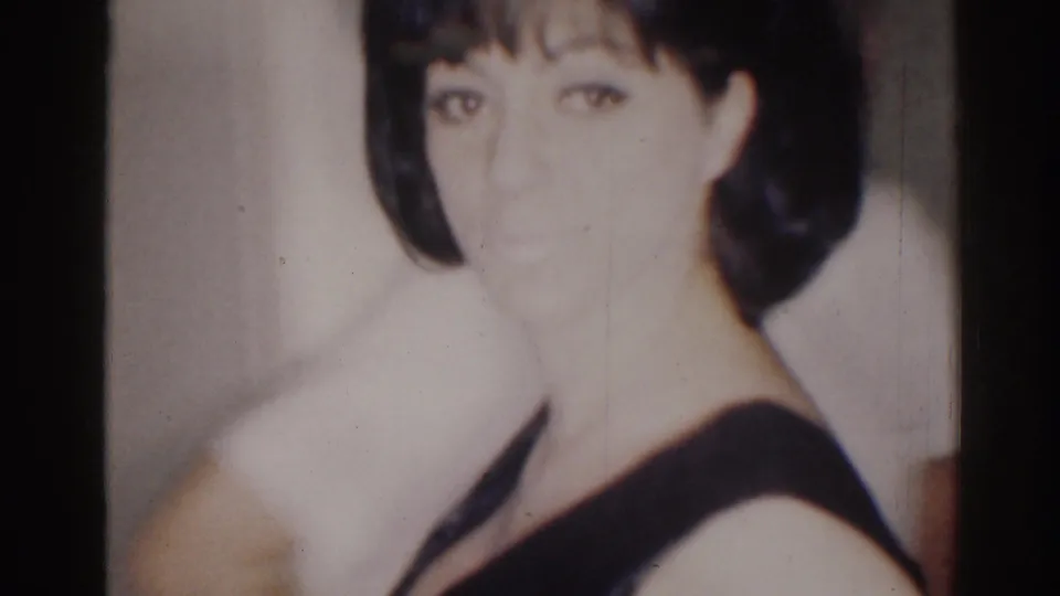 1967 Los Angeles 8mm frame of an adult woman in a dark sleeveless dress looking toward the camera in a private interior.