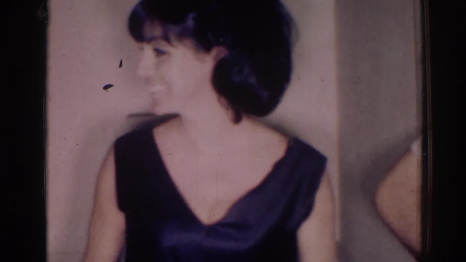1967 Los Angeles 8mm frame of an adult woman in a dark dress turning sideways with a smile inside a private room.