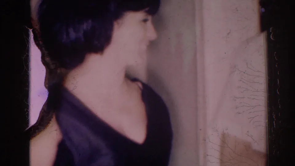 1967 Los Angeles home-movie frame showing a woman with a short dark bob in a dark dress turning inside a dim room.