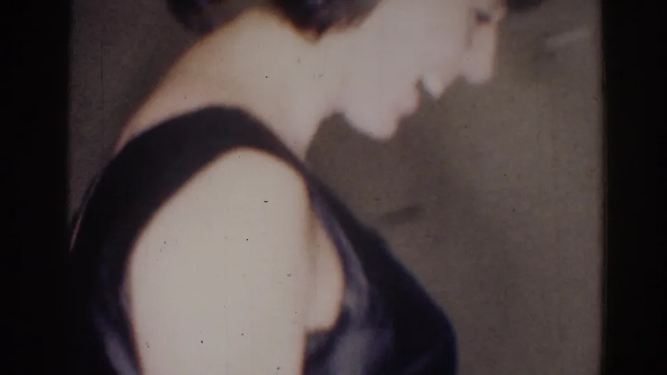 A woman in a dark sleeveless dress appears close to the camera in a dim 1967 Los Angeles interior.