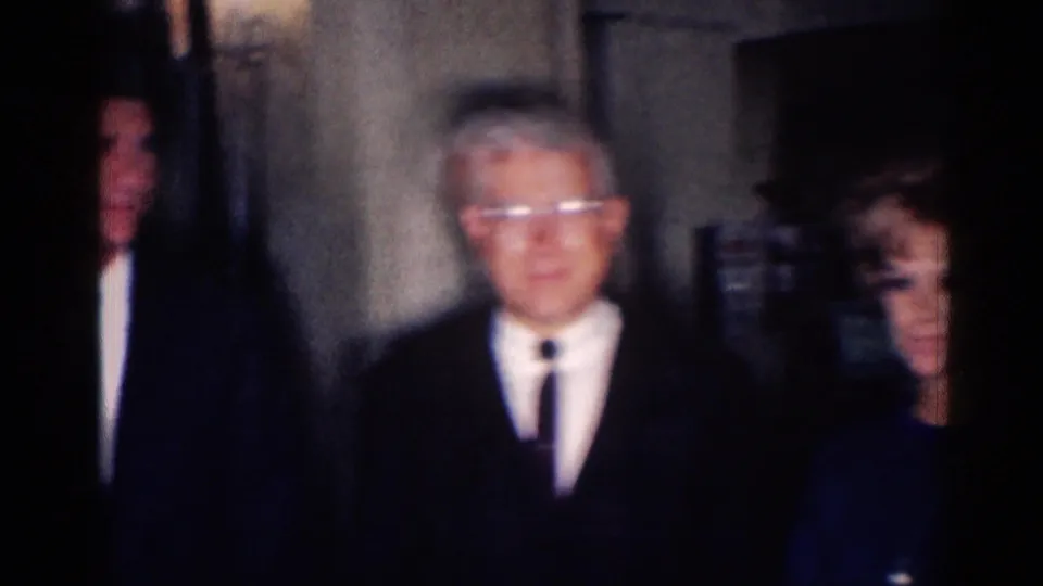 1967 Los Angeles frame with a suited man wearing glasses in a hallway, with other adults partly visible around him.