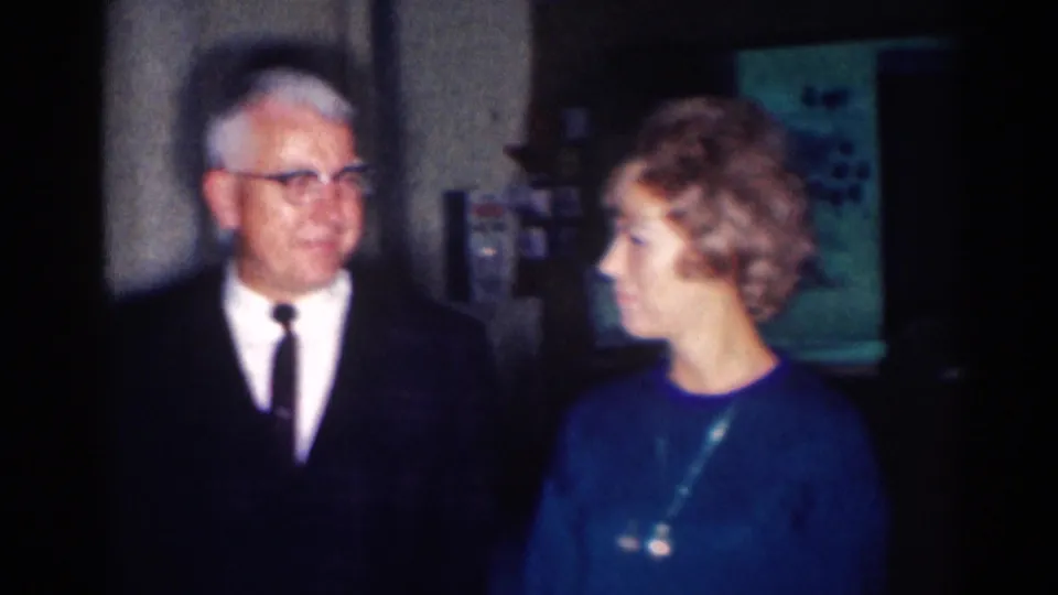 A suited man with glasses stands beside a woman in a blue dress in a 1967 Los Angeles home-movie interior.