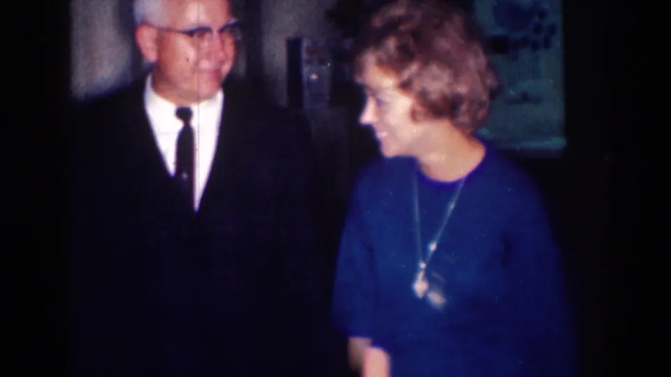 In 1967 Los Angeles home-movie footage, an older suited man with glasses stands near a woman in a blue dress indoors.