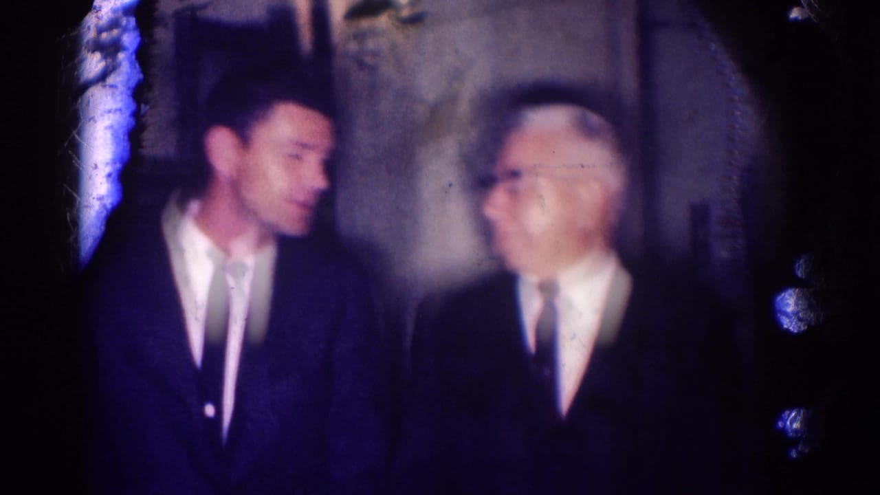 Younger man in a dark suit speaks with an older man wearing glasses indoors.