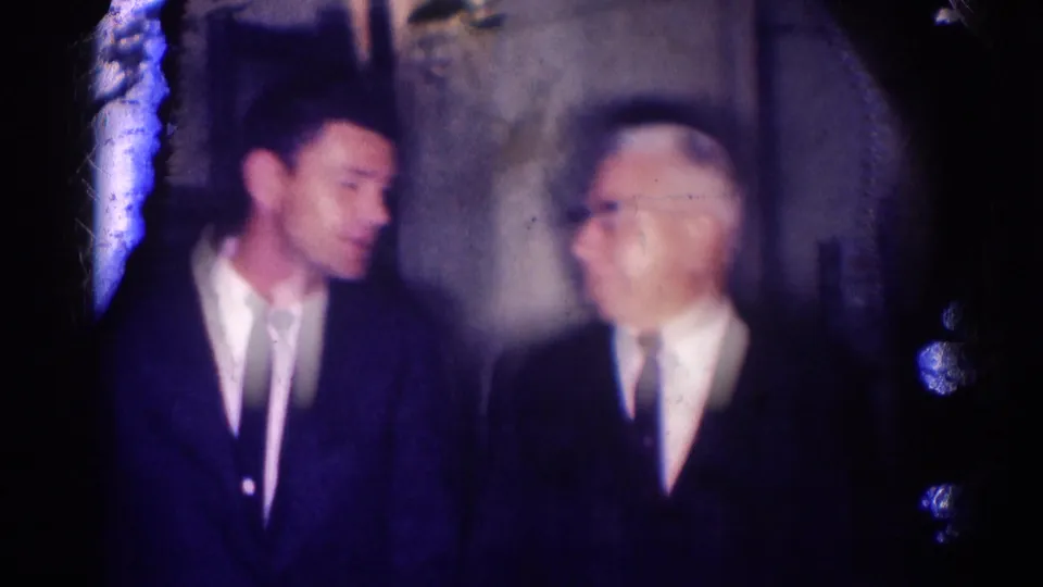 A younger suited man faces an older man with glasses during a dimly lit indoor conversation in 1967 Los Angeles footage.