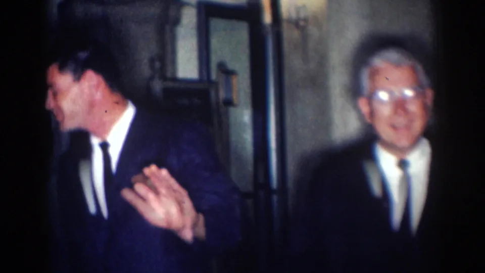 A man in a dark suit gestures with one hand as an older suited man stands in the dim 1967 Los Angeles interior scene.