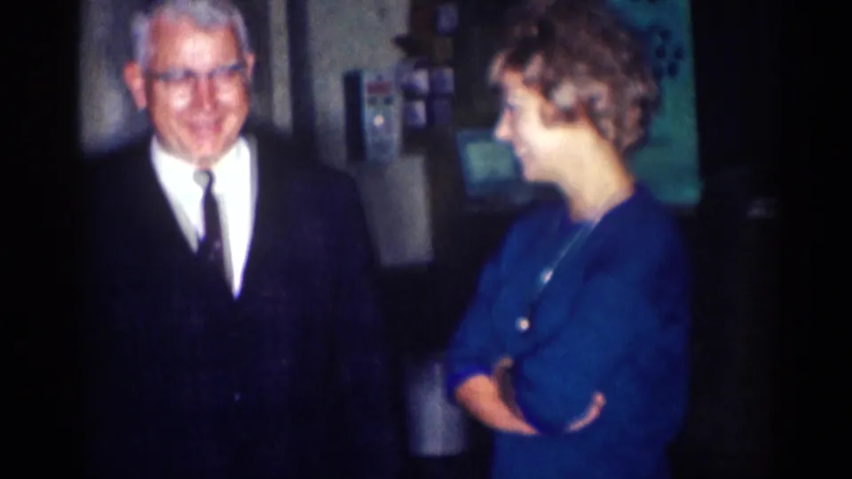 An older suited man smiles beside a woman in a blue dress with crossed arms in the 1967 Los Angeles interior footage.