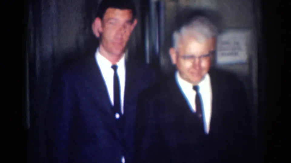 1967 Los Angeles 8mm frame showing two adult men in dark suits and narrow ties in a low-lit interior hallway.