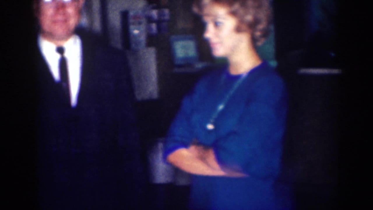 A suited man stands near a woman in a blue dress inside a dark room.