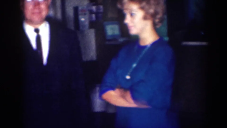 1967 Los Angeles 8mm frame with a suited man and a woman in a blue dress standing in a dim private interior.