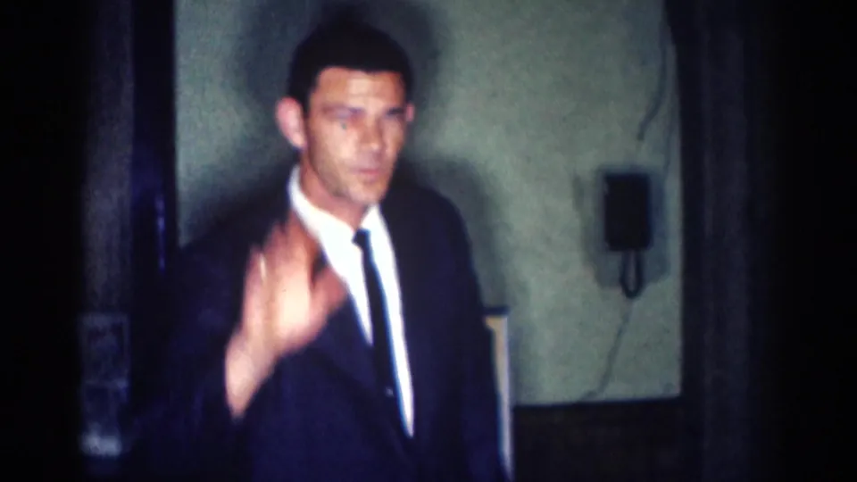 1967 Los Angeles 8mm frame showing an adult man in a dark suit raising his hand toward the camera in a dim interior.