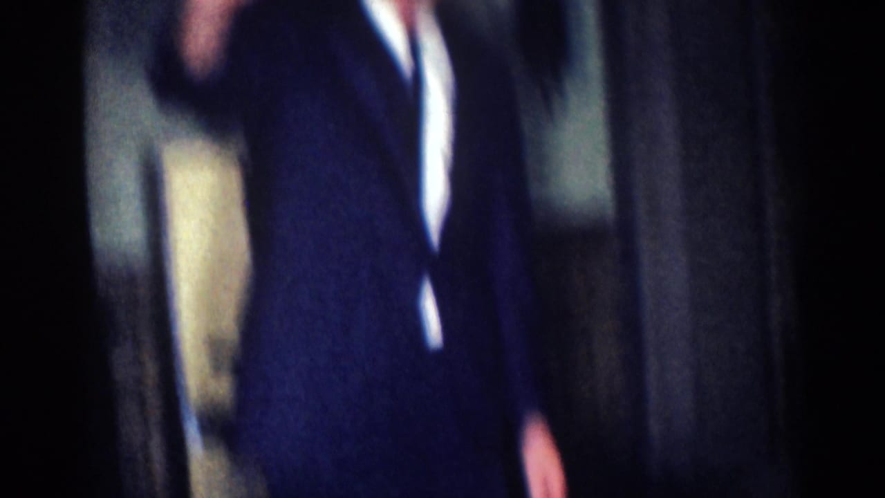 Man in a dark suit partly visible indoors with one hand raised near the camera.
