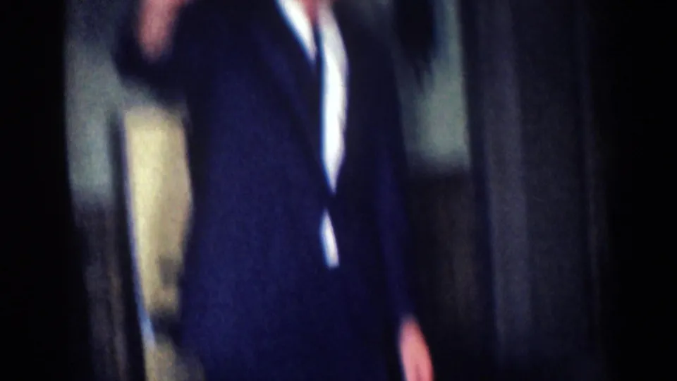 1967 8mm frame from Los Angeles metadata showing a suited man cropped at the head as he raises a hand in a dim interior.