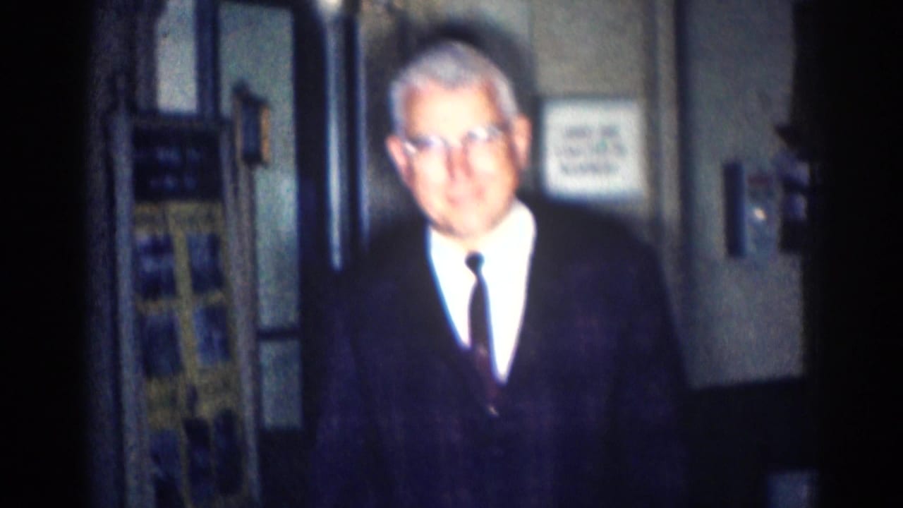 Older man with glasses in a dark suit stands indoors facing the camera.