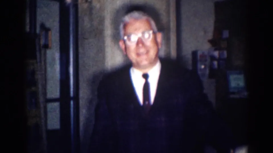 1967 8mm frame shows an older suited man with glasses centered in a dim Los Angeles interior, looking toward the camera.