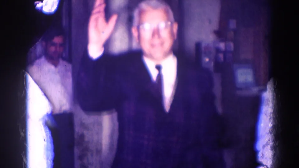 Older man with glasses waves toward the camera in a 1967 8mm Los Angeles interior, with another adult partly visible behind him.
