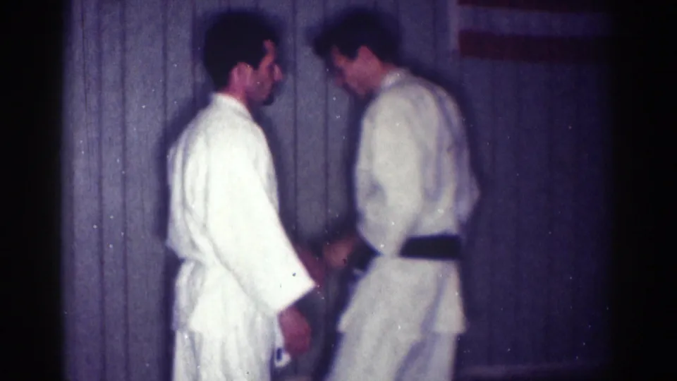 1967 Los Angeles home-movie frame of two adult men in white karate gi standing close during indoor martial arts practice.