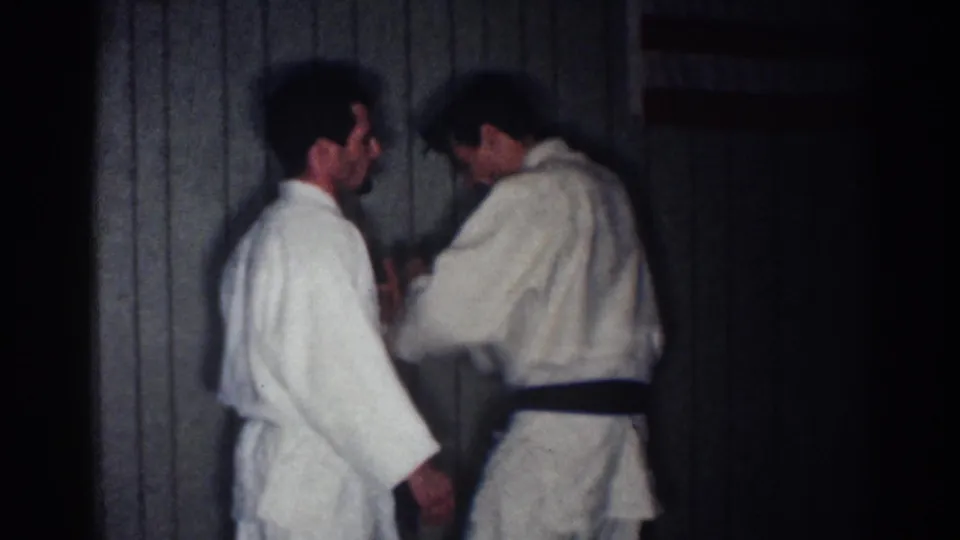 1967 Los Angeles home-movie frame showing a black-belt karate practitioner demonstrating close hand placement to another adult man.