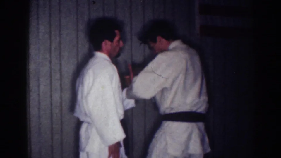 1967 Los Angeles home-movie frame of a black-belt practitioner showing close hand contact during a karate or self-defense lesson.