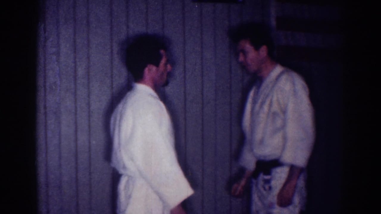 Two men in white martial arts uniforms face each other indoors near a paneled wall.