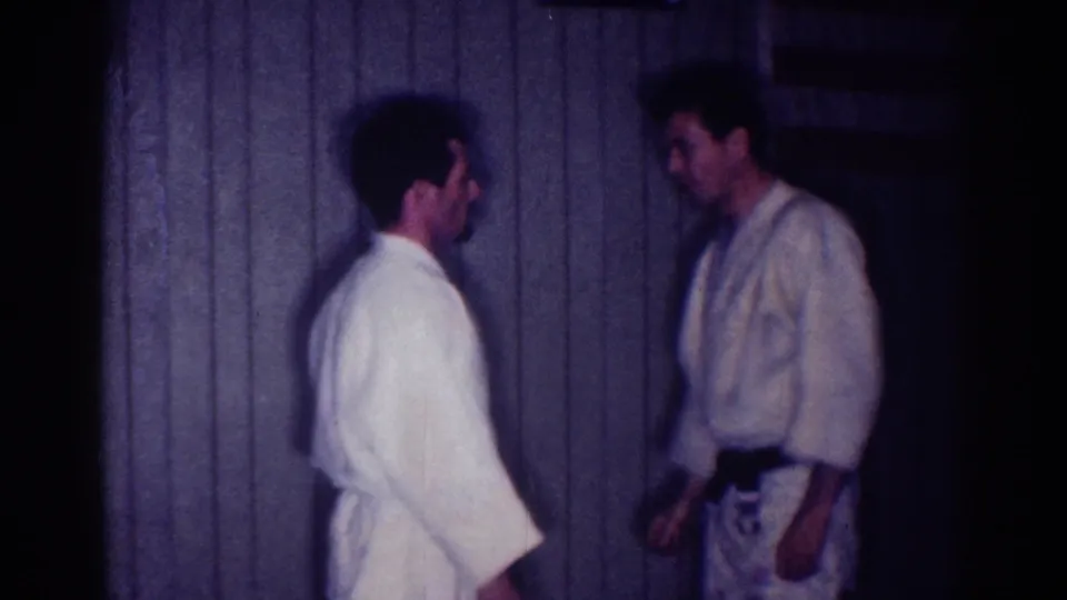 Two adult men in white martial arts uniforms stand facing each other in a dim Los Angeles interior from 1967 home-movie footage.