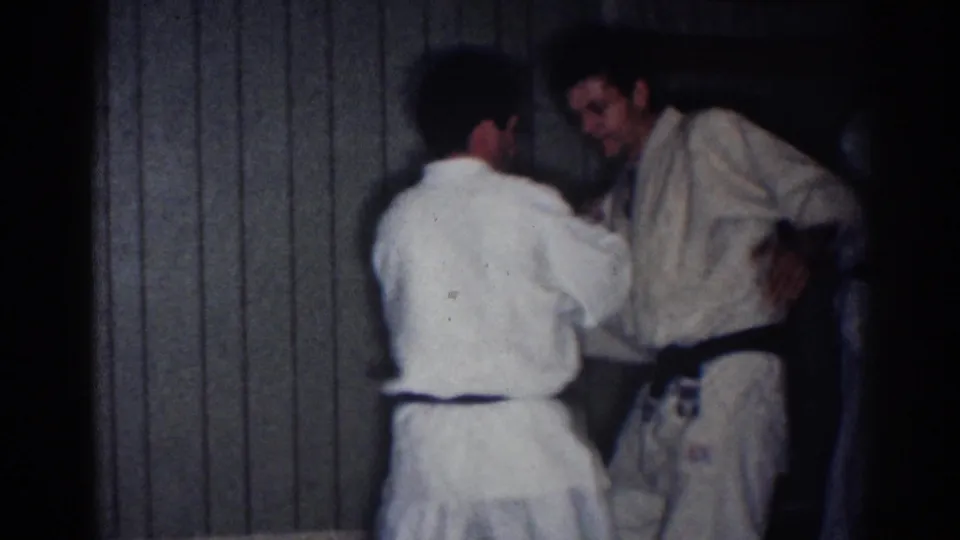 Two adult men in white martial arts uniforms practice a close-range technique in the dim paneled interior of the 1967 Los Angeles clip.