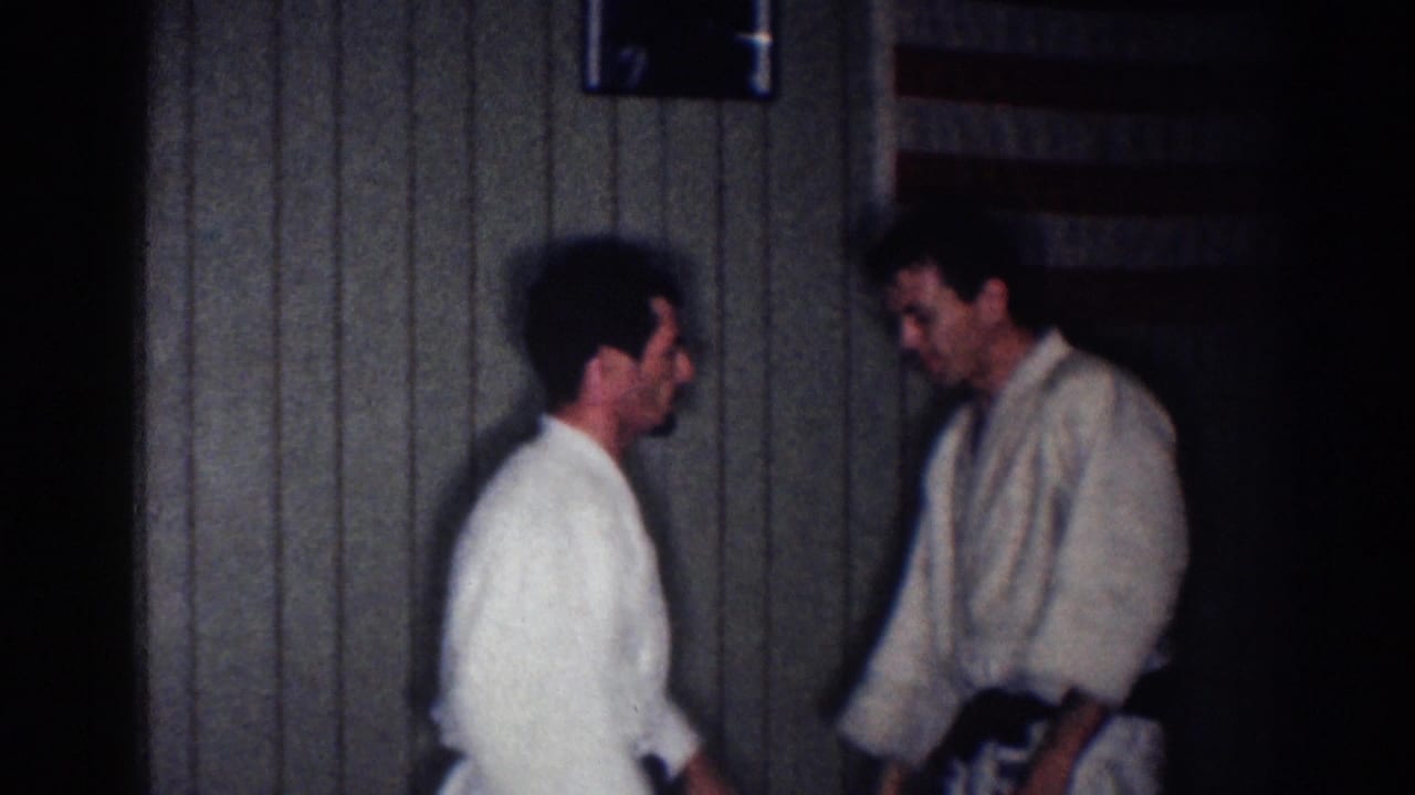1967 Los Angeles home-movie frame showing two men in gi uniforms facing each other in a wood-paneled dojo interior.