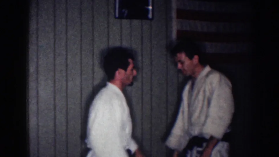 1967 Los Angeles home-movie frame showing two men in gi uniforms facing each other in a wood-paneled dojo interior.