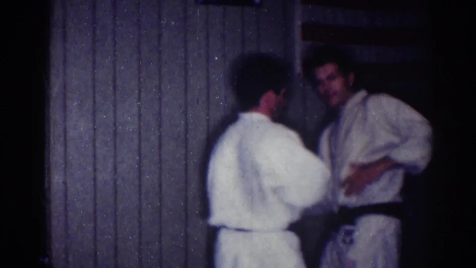 Two adult men in karate uniforms pause near a paneled wall and American flag in the 1967 Los Angeles dojo footage.