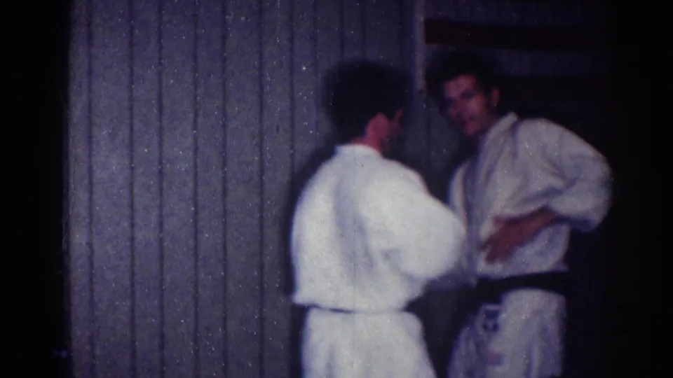 Home-movie frame of two men in karate uniforms standing close during practice inside a Los Angeles dojo in 1967.