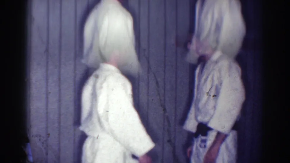The white-belt practitioner faces a black-belt man in a dim 1967 Los Angeles karate dojo interior.