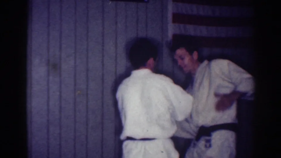 In a 1967 Los Angeles home-movie frame, two men in white gi practice a close hand-grab technique indoors.