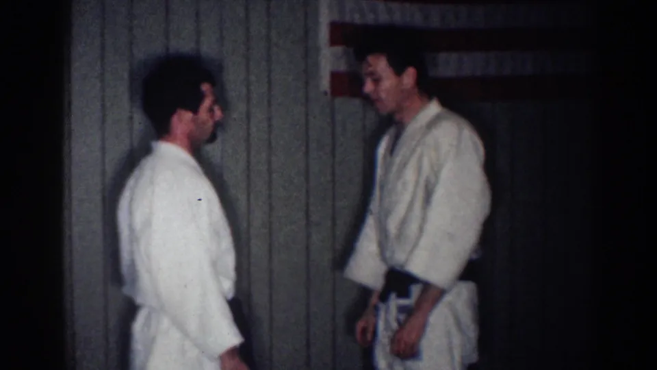 Two adult men in white gi face one another inside a dim Los Angeles training room with an American flag behind them.