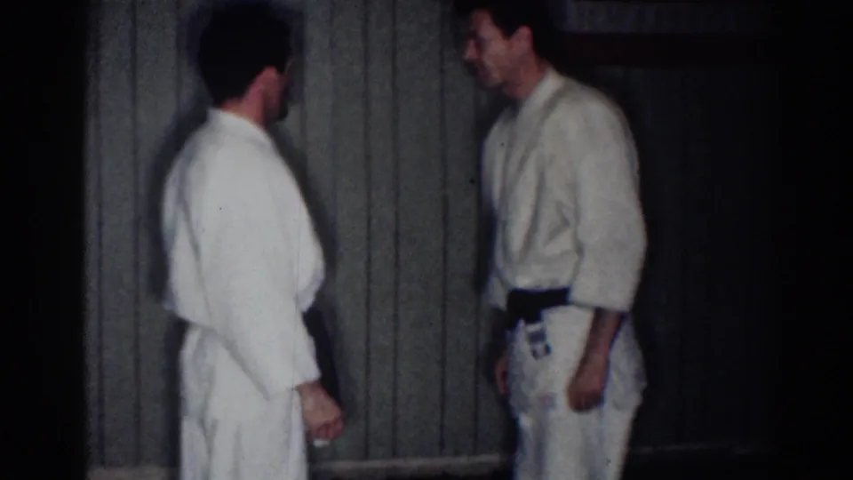 Two men in white gi stand facing each other in the dim indoor practice space, with wood paneling and a dark doorway visible.