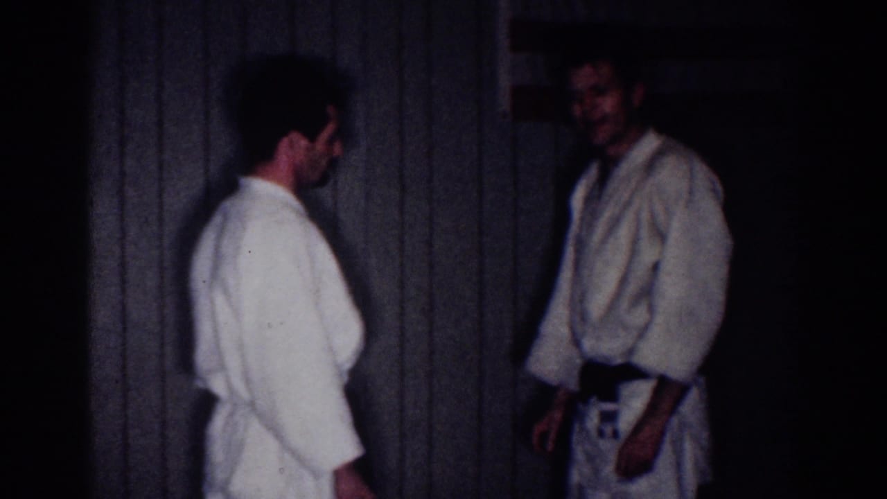 Two men in white martial arts gis stand in a dim paneled room.