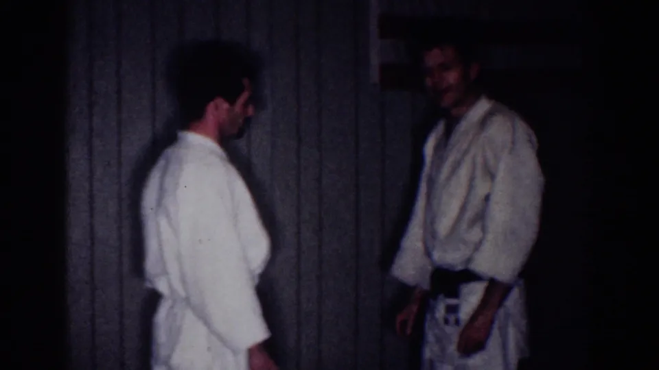 Two adult men in white martial arts gis face each other in a dim Los Angeles indoor room, filmed on 8mm in 1967.