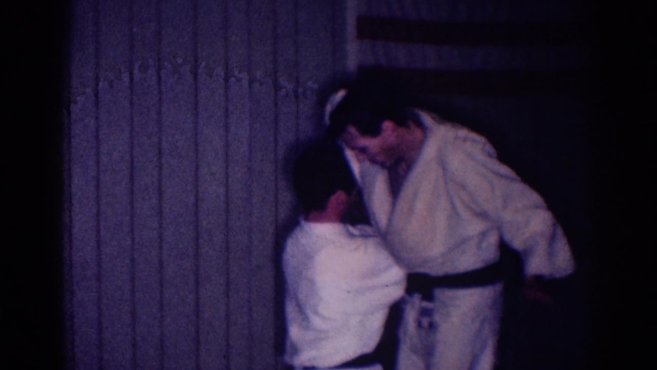 One man in a white gi leans toward another during a close martial arts hold.