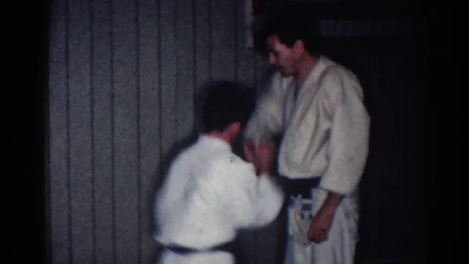 One adult man crouches or kneels while practicing an arm technique with a standing man in a white gi and black belt.