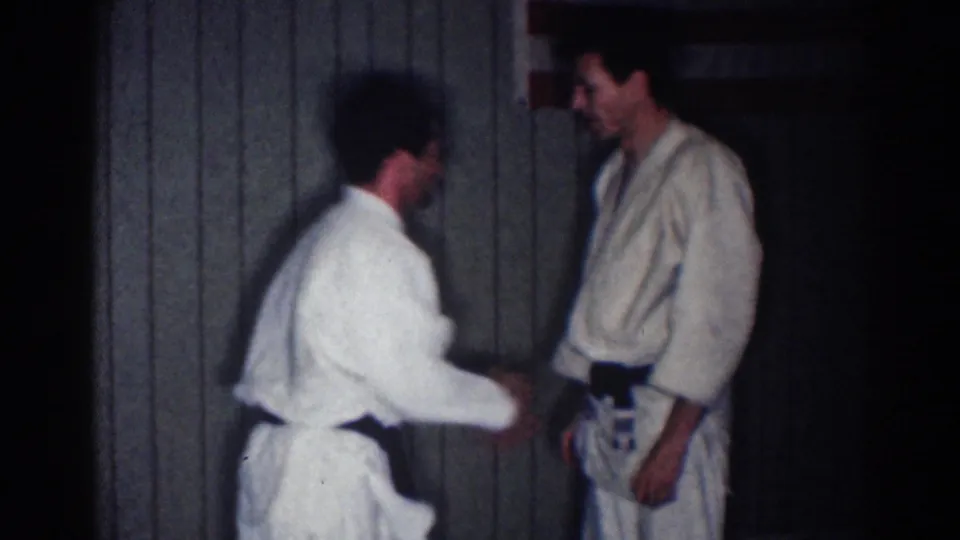Two adult men in white martial arts uniforms practice a hand or wrist hold in a dim indoor Los Angeles setting, based on the clip metadata.