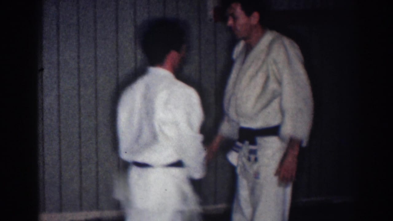 Two athletes in white judo uniforms face each other near a wood-paneled wall.