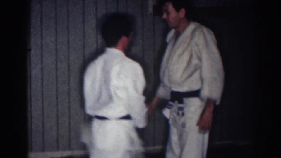 1967 Los Angeles home-movie frame showing two judo athletes in white gi and dark belts facing one another indoors.