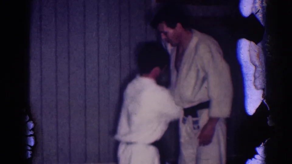 Dim 1967 Los Angeles 8mm frame showing two athletes in gi demonstrating a close hold near a wood-paneled wall.