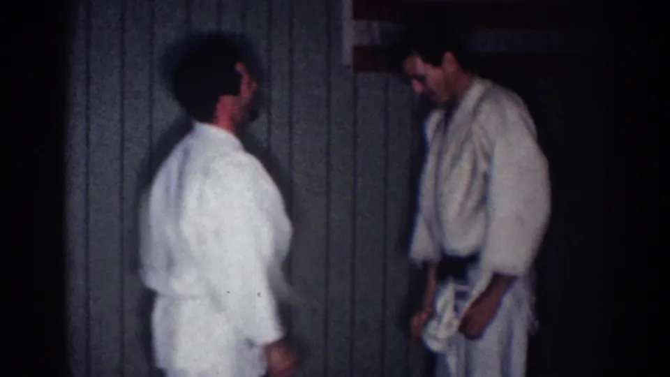 1967 Los Angeles home-movie frame with two judo athletes in white gi facing each other against a plain indoor wall.