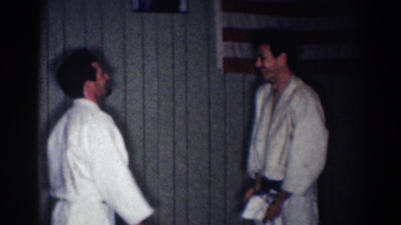 Two adult men in white gi uniforms face each other in a dim Los Angeles interior, filmed as 1967 8mm home-movie footage.