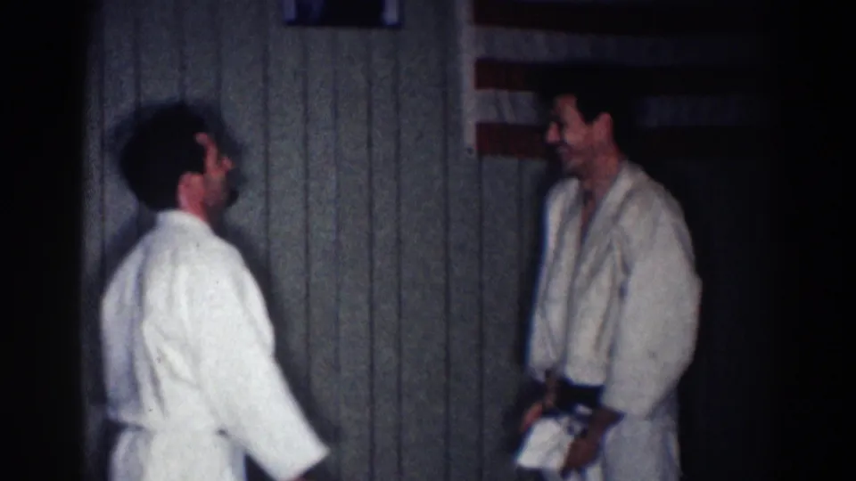 Two adult men in white gi uniforms face each other in a dim Los Angeles interior, filmed as 1967 8mm home-movie footage.