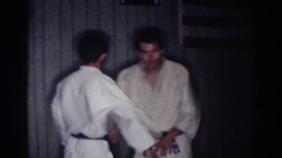 One adult man in a white gi reaches toward another during a martial arts practice exchange inside a dim 1967 Los Angeles room.