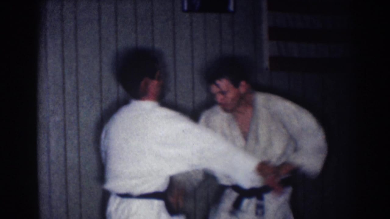 Two adult men in white gi uniforms practice a close-contact martial arts technique in soft, grainy 1967 Los Angeles footage.