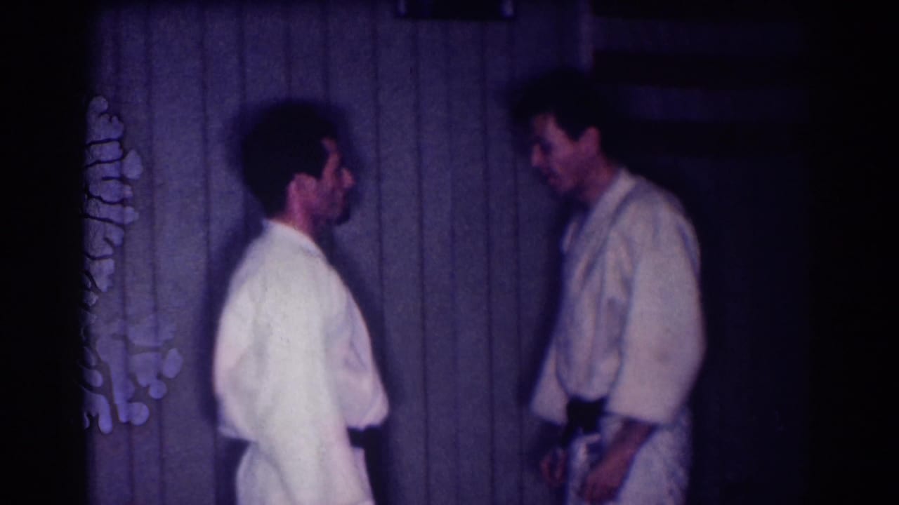 Two adult men in white gi uniforms stand apart after a practice exchange in a dim 1967 Los Angeles home-movie frame.