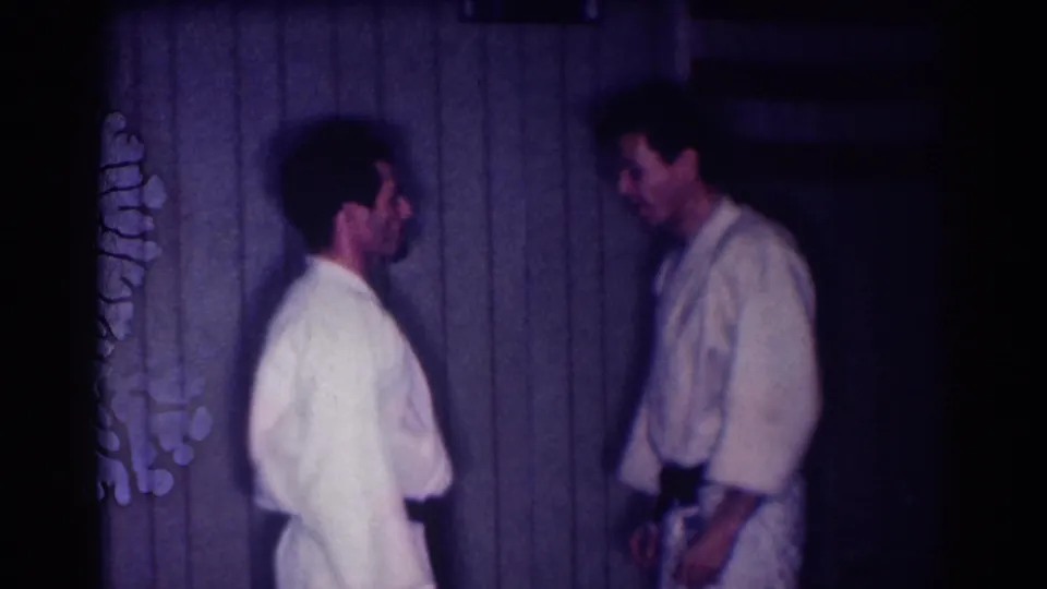 Two adult men in white gi uniforms stand apart after a practice exchange in a dim 1967 Los Angeles home-movie frame.